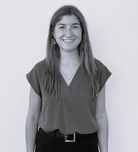 Nora Balboni, with long hair, stands facing the camera, smiling. She is wearing a short-sleeve top with a V-neck, a necklace, a belt, and dark pants. The background is plain. The image is in black and white.