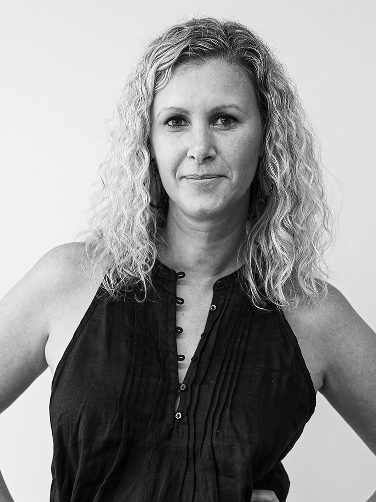 Black and white portrait of Nadia Greatbatch with curly, shoulder-length hair. She is wearing a sleeveless, button-up top and has a neutral expression. The background is plain, and she has one hand resting on her hip.