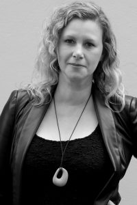 A black and white portrait of Nadia Greatbatch, a woman with wavy, shoulder-length hair. She is wearing a textured black top, a leather jacket, and a pendant necklace. She stands against a plain background, looking directly at the camera with a neutral expression.