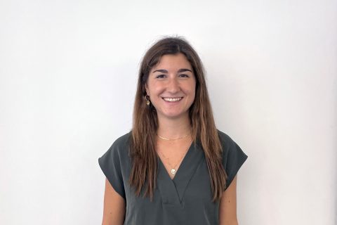 A woman with long, straight brown hair is standing against a plain white background. She is wearing a dark green short-sleeve blouse and smiling at the camera. Her name is Nora Balboni, and she has a gold necklace and small hoop earrings.