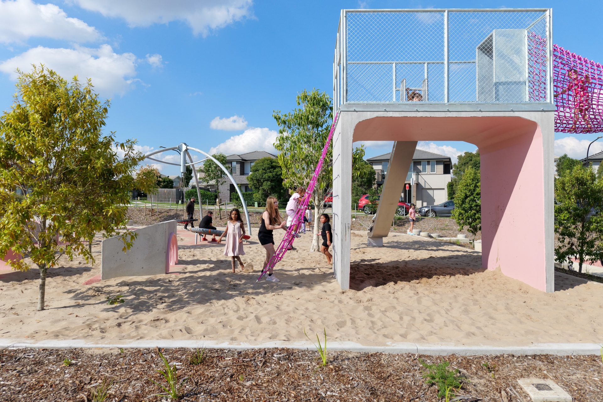 A group of children and an adult are playing in a sandy playground with various climbing structures, including a pink climbing net. At Galungara Park, the playground is surrounded by trees and residential buildings. The sky is clear with a few clouds.