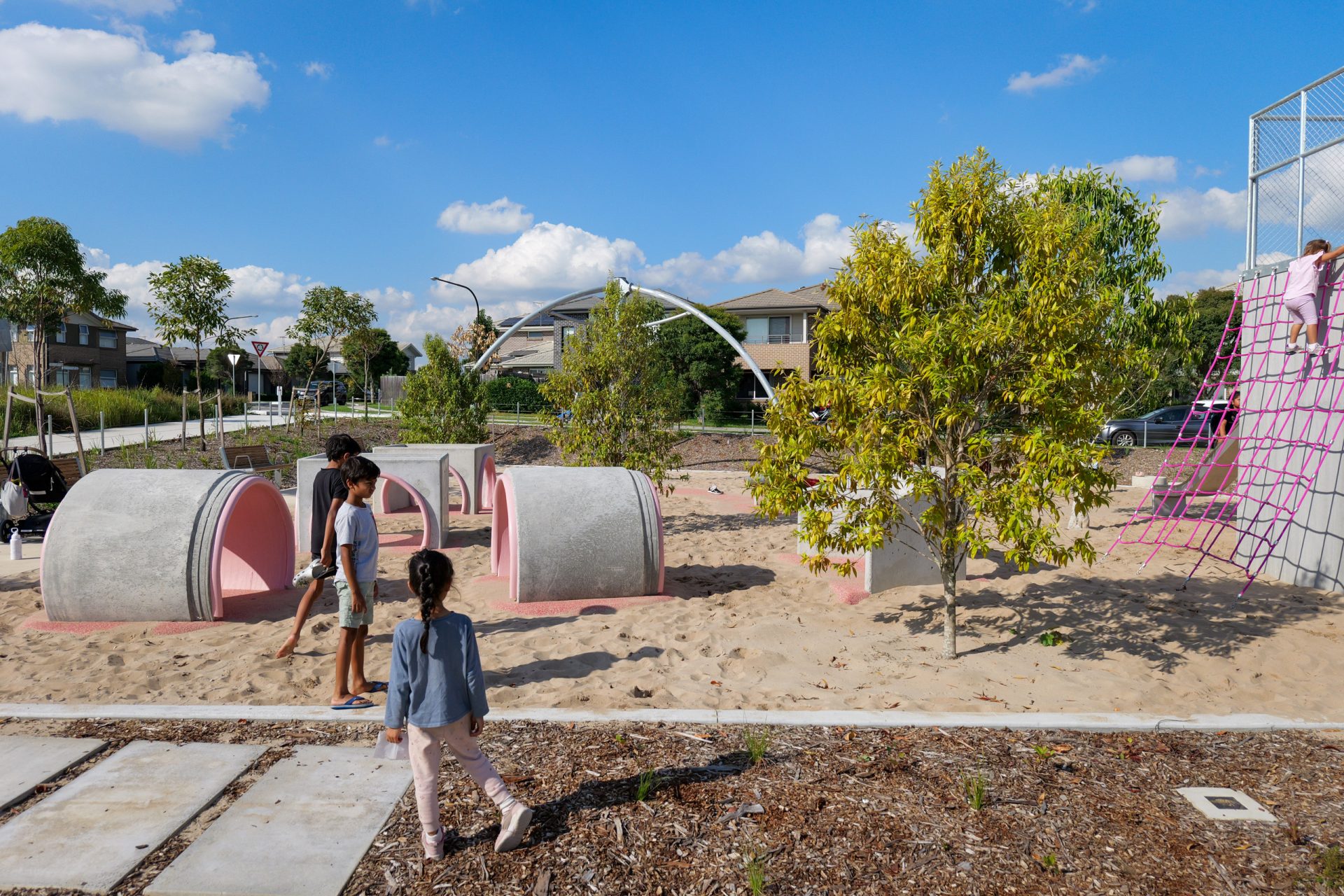 At Galungara Park, two children walk on a sandy surface. The playground features concrete tunnel structures, climbing nets, and surrounding trees. In the background, residential houses and a clear blue sky with scattered clouds are visible.