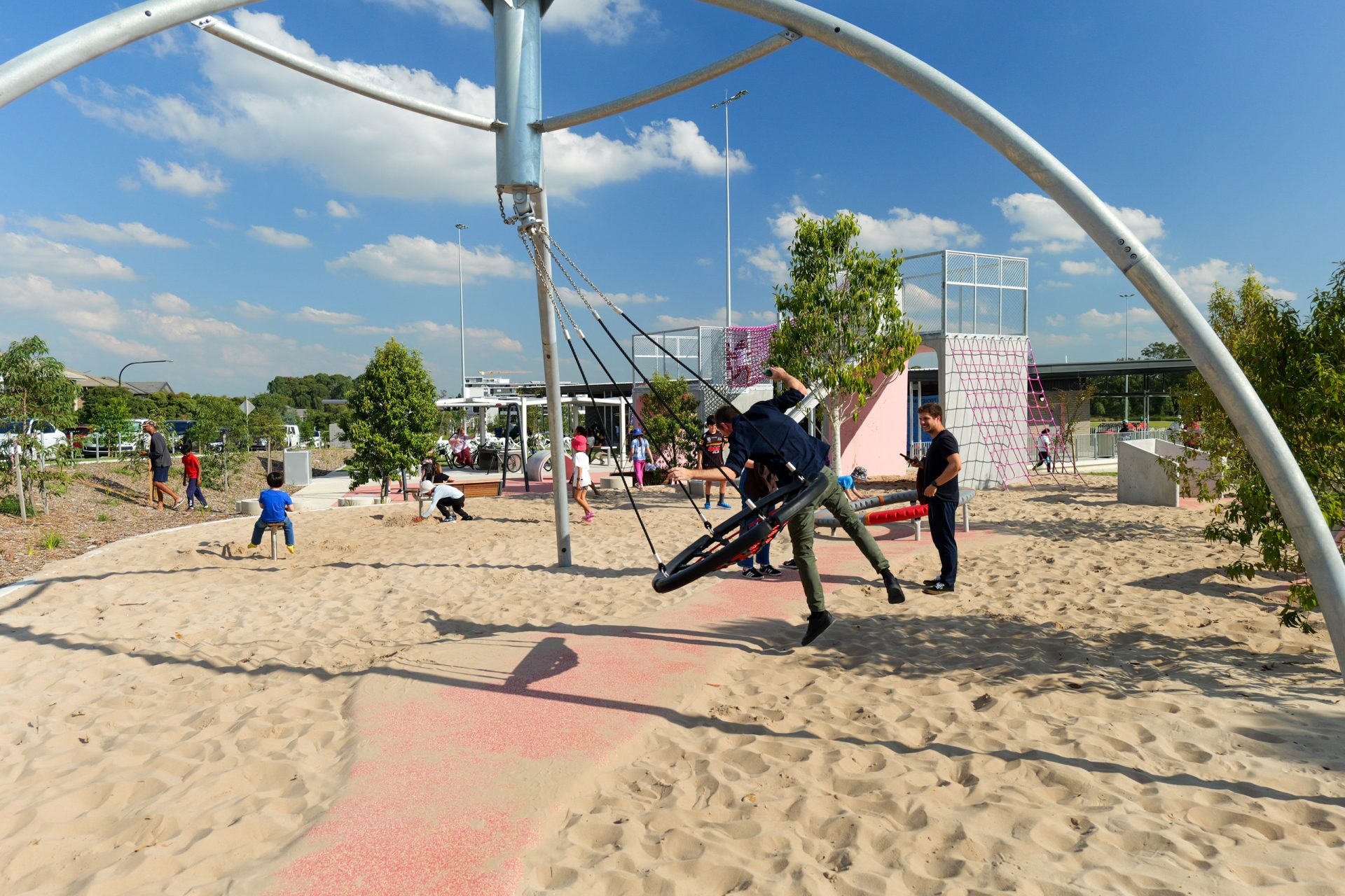 Children and adults enjoy a sunny day at Galungara Park's modern playground. A large, circular swing with multiple seats is the focal point, holding several kids. Other children play in the sandy area and climb on a pink, geometric structure. Trees and open skies complete the scene.