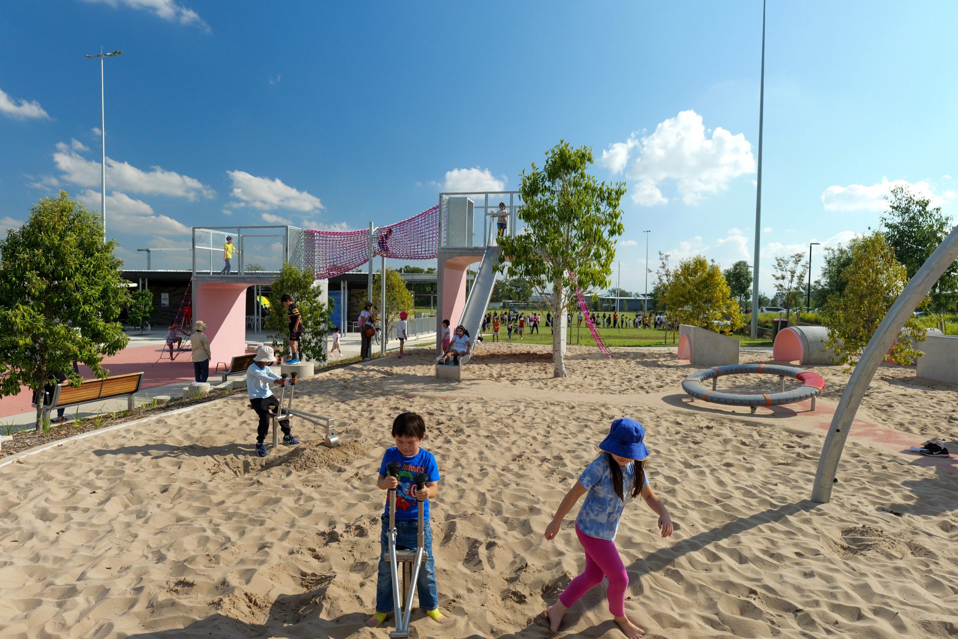 Children play in the sandy area at Galungara Park on a sunny day. One child rides a scooter, another digs with a shovel, and a third walks by. The playground features slides, climbing structures, trees, and benches, with parents and more children in the background.