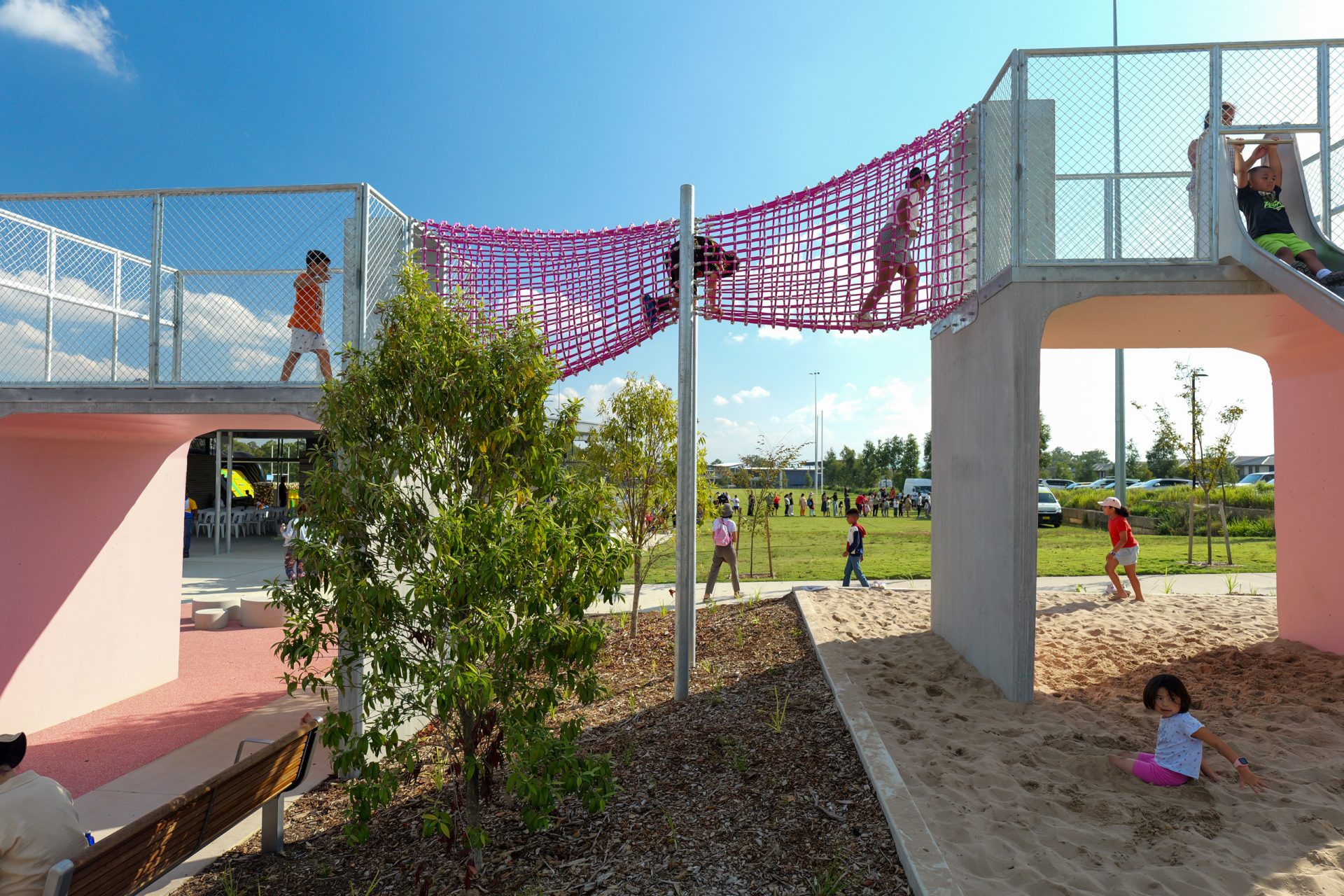 Children play on the Galungara Park playground with elevated walkways covered by pink netting. One child climbs the netting, while others explore the sandy area below. A bench and leafy bushes are in the foreground, and green grass, trees, and people are in the background.