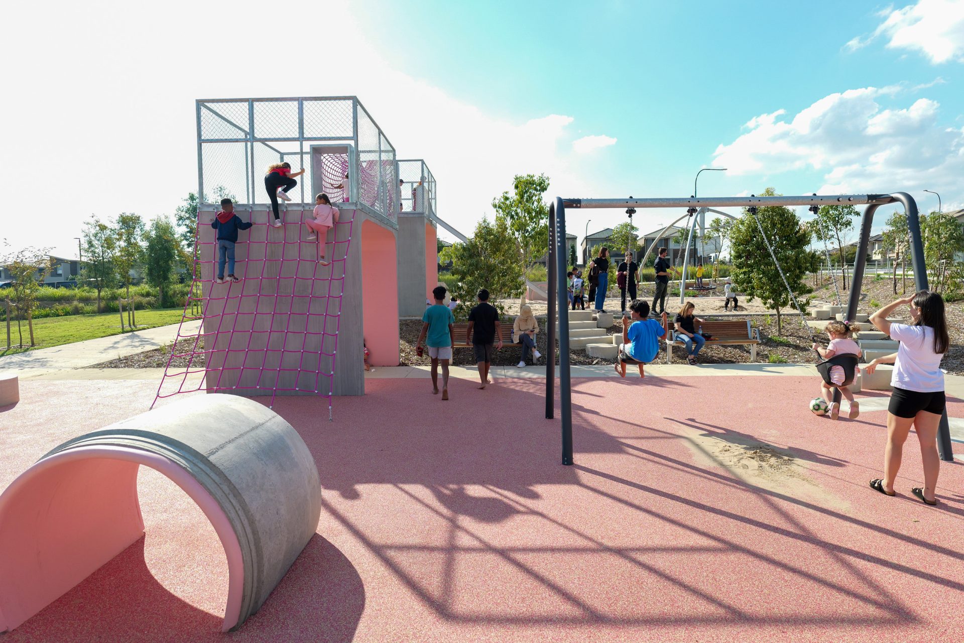 A sunny day at Galungara Park's vibrant playground features children and adults enjoying themselves. A climbing structure with a rope net and ladder stands on the left, a swing set on the right, and a large concrete pipe in the foreground. Trees and a grassy area provide a serene backdrop.