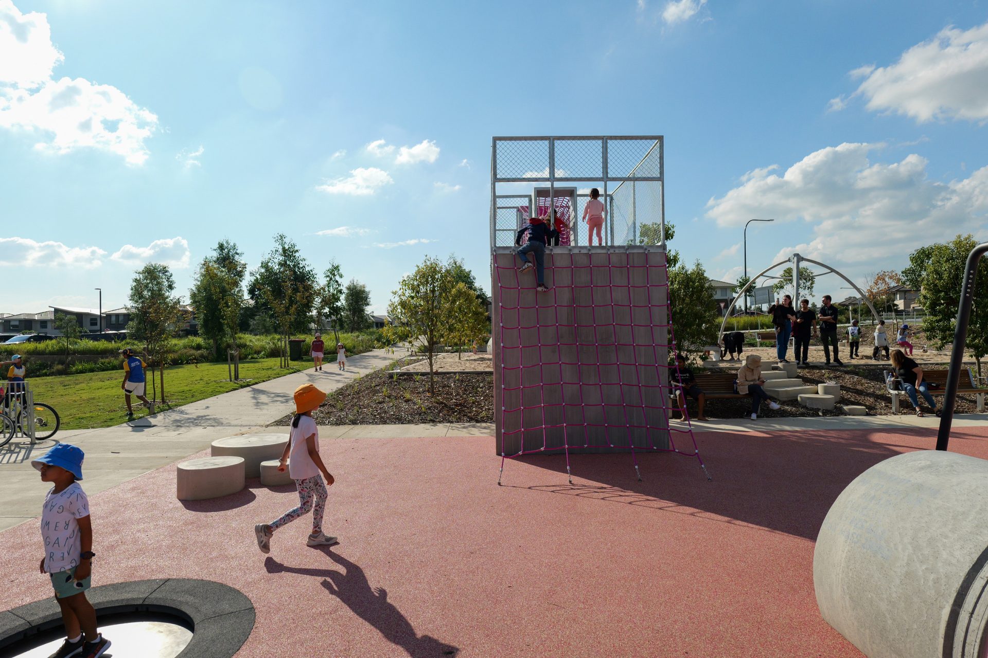 Children play at Galungara Park, a colorful, outdoor playground featuring climbing structures and open spaces. One child walks near the pink and red rubber surfacing. Several children climb a net to reach a raised platform. Adults stand in the background near benches and trees under a blue sky.