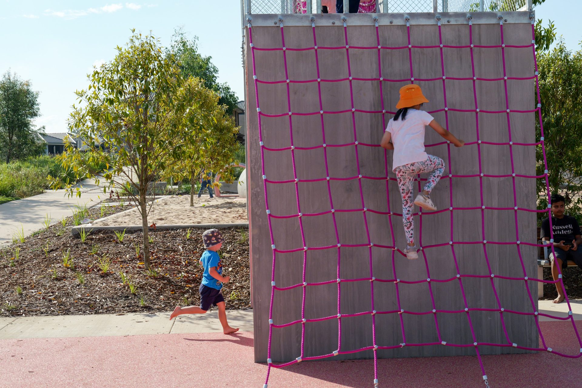 At Galungara Park, a child wearing a bright orange hat and patterned pants climbs a pink rope wall at the playground. Another child in a blue shirt and patterned shorts runs past the wall. Trees and a pathway are visible in the background, while other children play nearby.