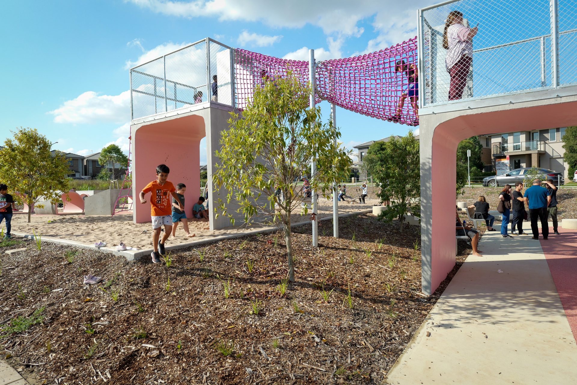 Children and adults interact in the vibrant Galungara Park playground. Some children climb a pink net bridge connecting two elevated platforms, while others explore the ground below. The area is adorned with trees and mulch, and there are residential houses in the background.