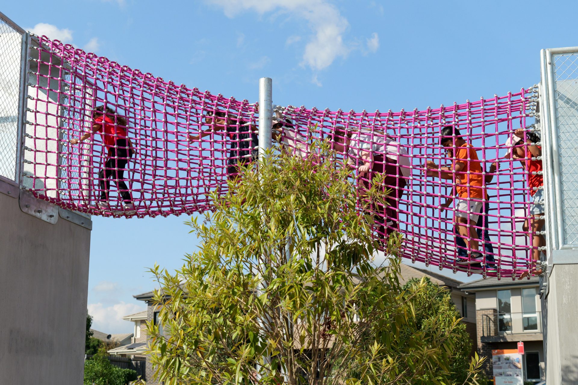 Children are climbing and playing on a large pink net structure suspended between two platforms at Galungara Park. A tall bush is visible in the foreground, with some buildings and a clear blue sky in the background of this lively playground.