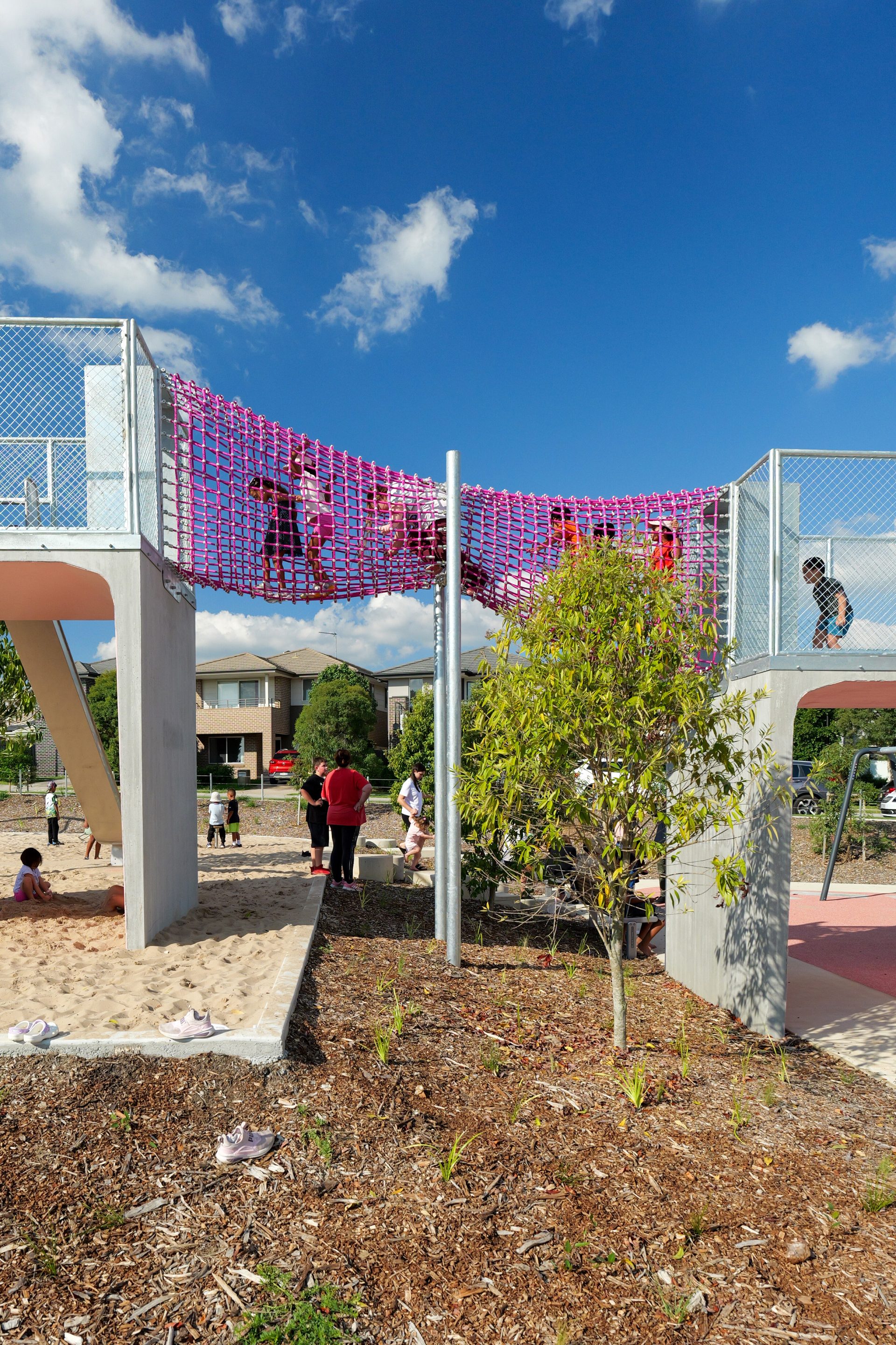 Children play on a high netted bridge at the Galungara Park playground, with a sand area beneath them. The playground is surrounded by plants, and modern residential buildings form the backdrop. The sky is clear with a few scattered clouds.