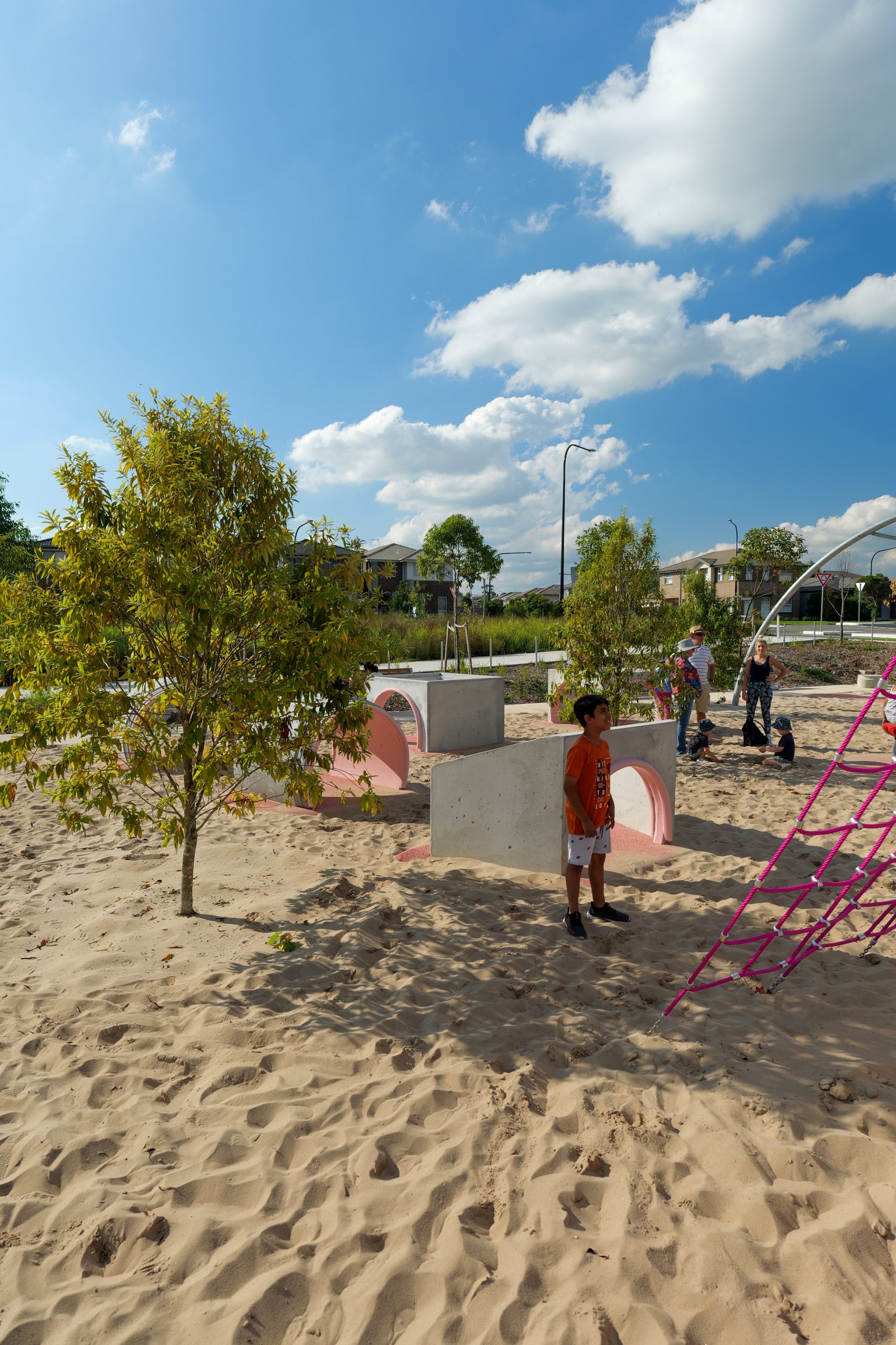 A child in an orange shirt stands on the sandy ground of Galungara Park's playground near a play structure with pink climbing nets, while adults and other children interact with the environment. Trees are scattered around, and houses and a clear blue sky with clouds are visible in the background.