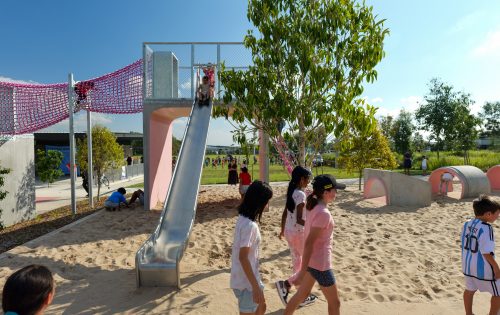 Children are playing in a sunny outdoor playground at Galungara Park. Some kids are using a metal slide, while others walk on the sandy ground or explore tunnel structures. A tree provides some shade, and various playground equipment is scattered in the background.