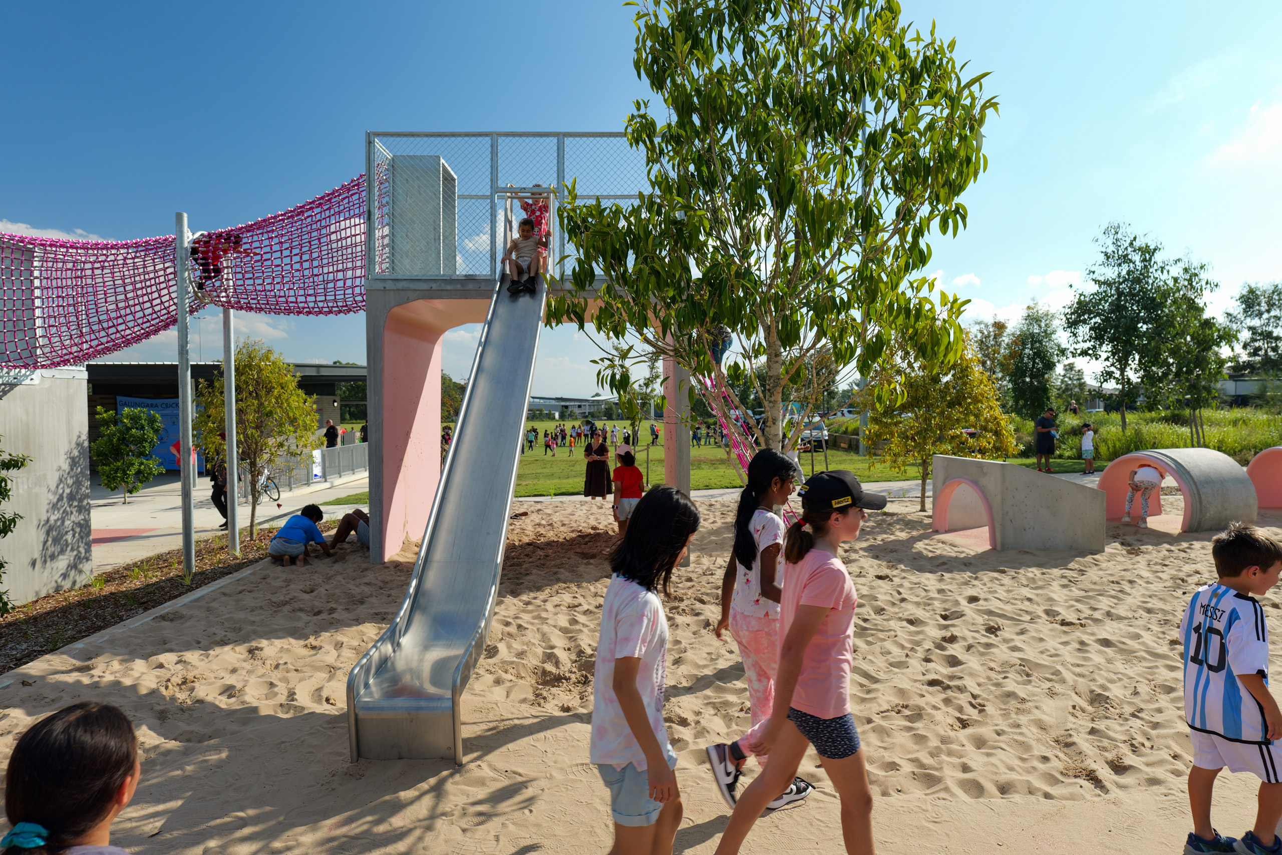 Children are playing in a sunny outdoor playground at Galungara Park. Some kids are using a metal slide, while others walk on the sandy ground or explore tunnel structures. A tree provides some shade, and various playground equipment is scattered in the background.