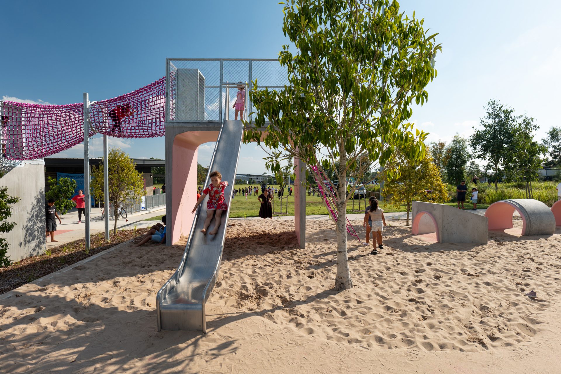 Galungara Park's playground features a slide, climbing net, and tunnels. Several children are happily playing, with one child sliding down. Adults stand nearby under clear skies. A tree provides some shade, and there's plenty of sand covering the ground. Nearby buildings and lush greenery complete the scene.