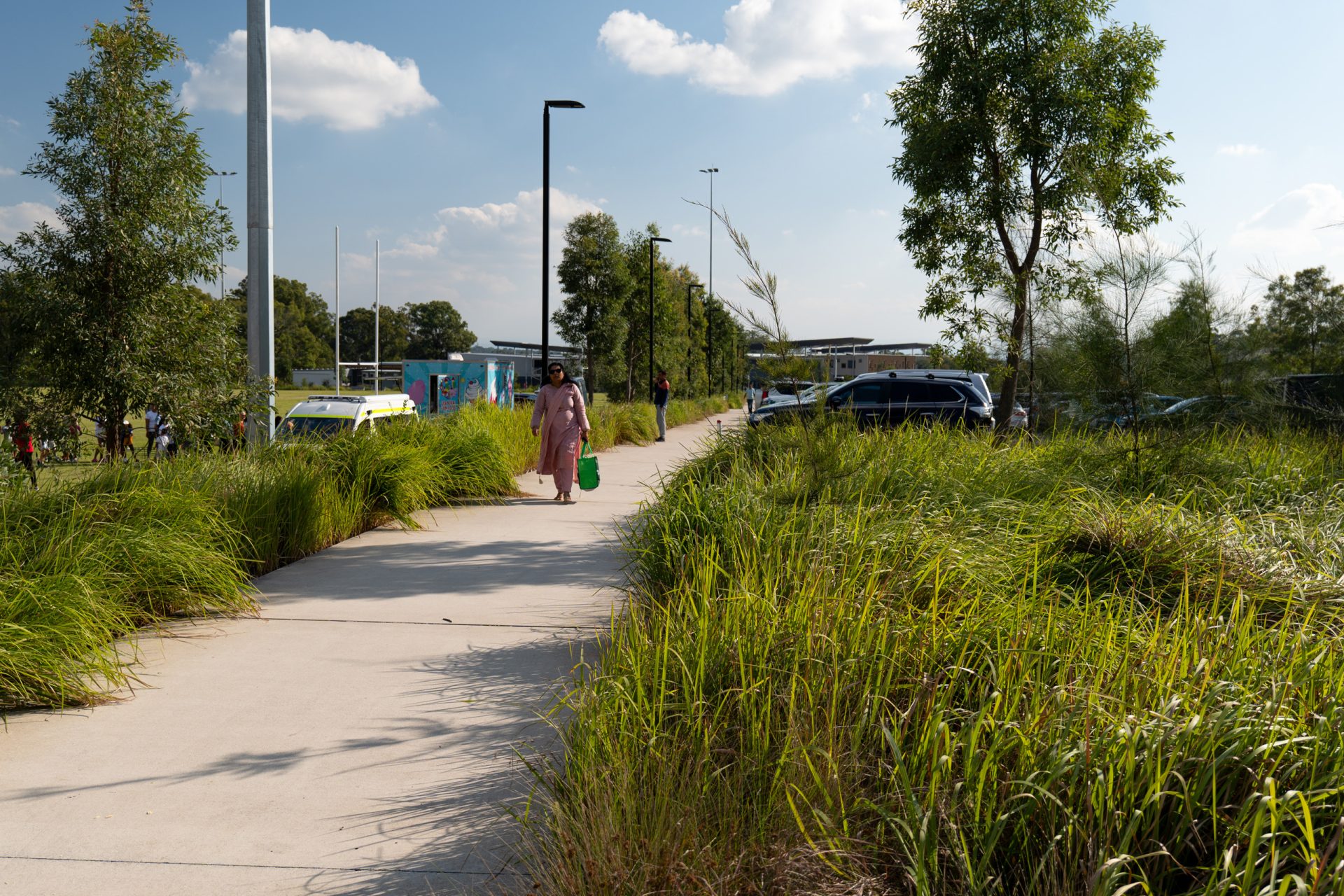 A person wearing a long, light-colored dress walks along a paved path surrounded by tall grass and trees under a clear sky. The path curves near Galungara Park's playground, with a parking lot and cars visible to the right, while more trees and a few people are seen in the background.