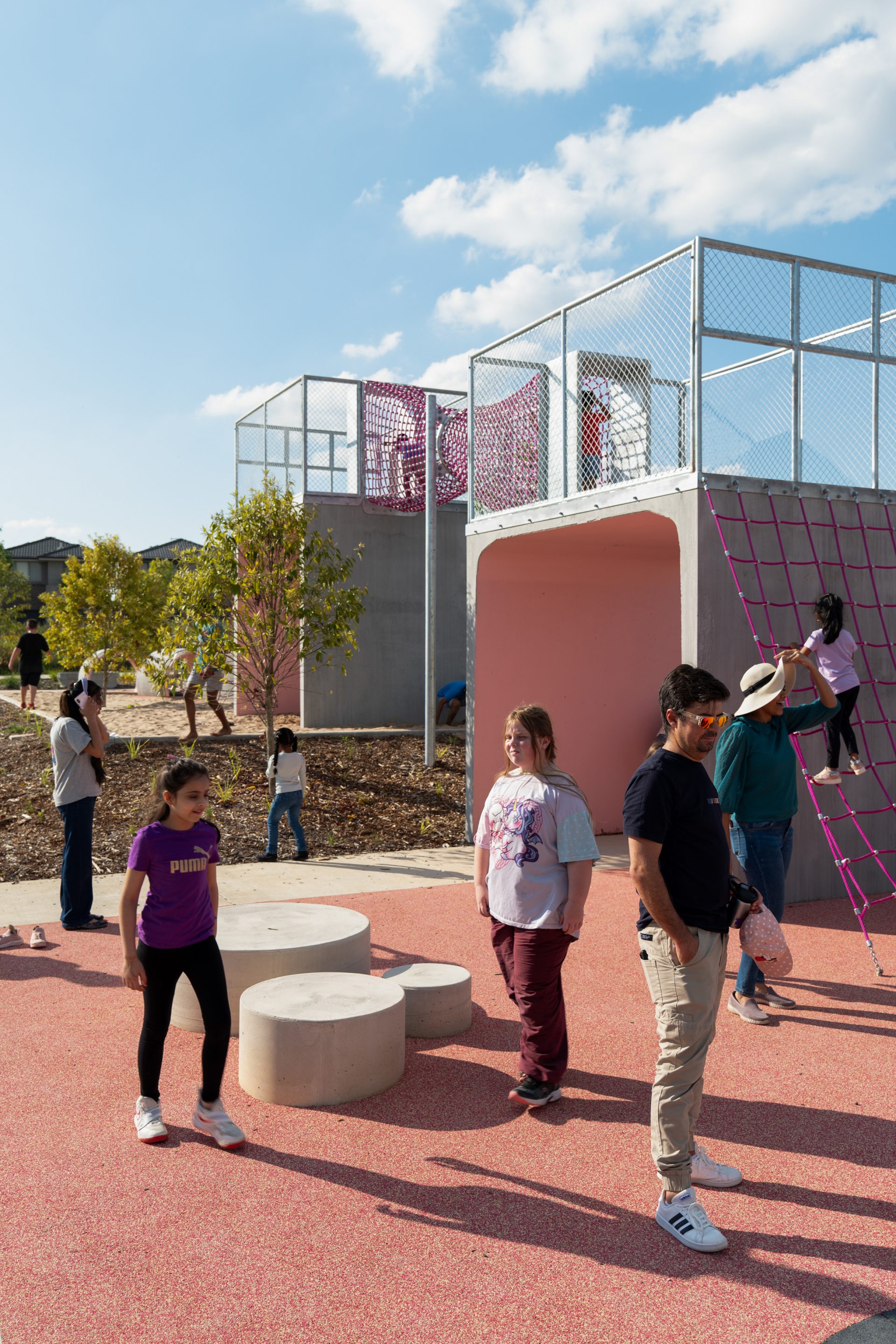 At Galungara Park, children and adults engage in various activities. Some kids climb a rope net on a pink structure, while others play on the ground. The playground features cement blocks, trees, and an open sky with scattered clouds in the background.