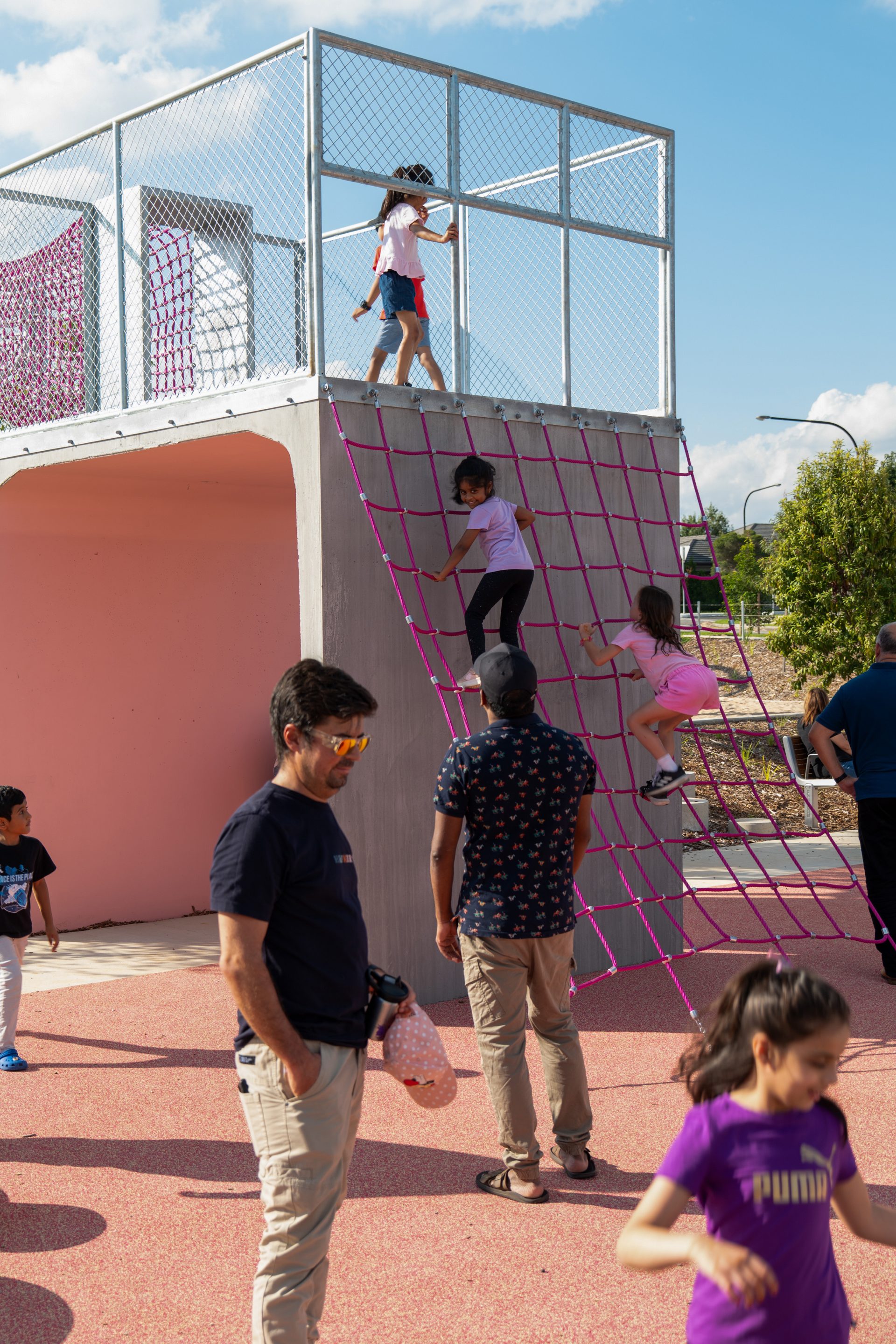 Children are playing on a playground at Galungara Park. Two girls climb a net on a pink structure while another girl stands at the top. Several adults and other children are on the ground engaging in various activities. The scene is bright and sunny with trees in the background.