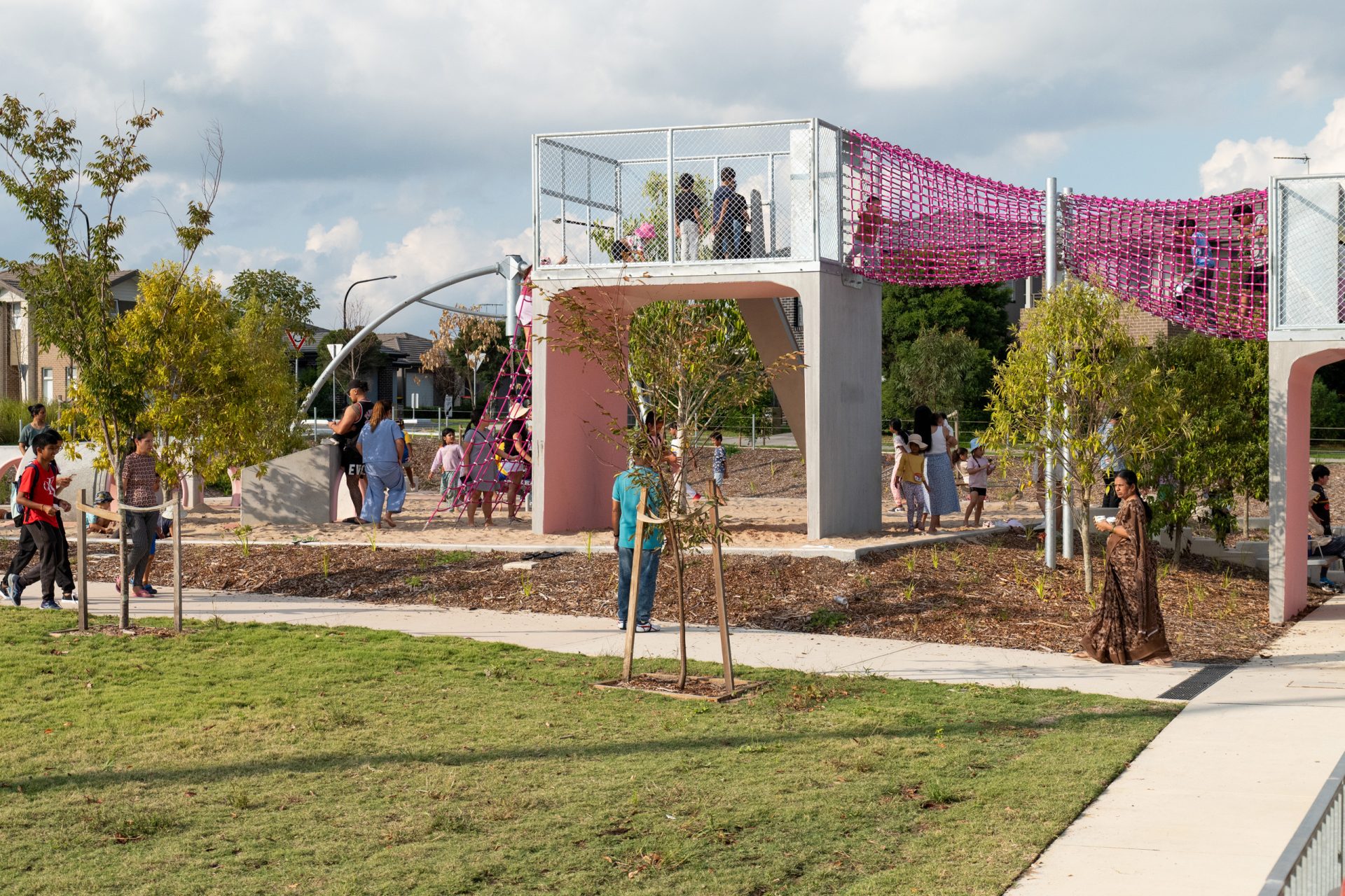 A diverse group of people enjoy a sunny day at Galungara Park. Some are climbing and walking on a modern playground with netted areas, while others walk and chat nearby on paths. The park is surrounded by trees and green grass, and the sky is partly cloudy.