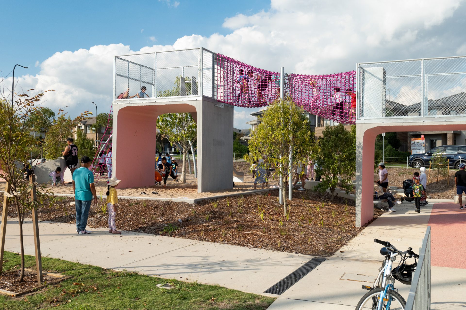 A vibrant scene at Galungara Park features a modern playground with elevated concrete structures connected by a pink net tunnel. Children gleefully climb and play, while adults relax nearby. A bicycle is parked in the foreground amidst trees and pathways that encircle the lively area.