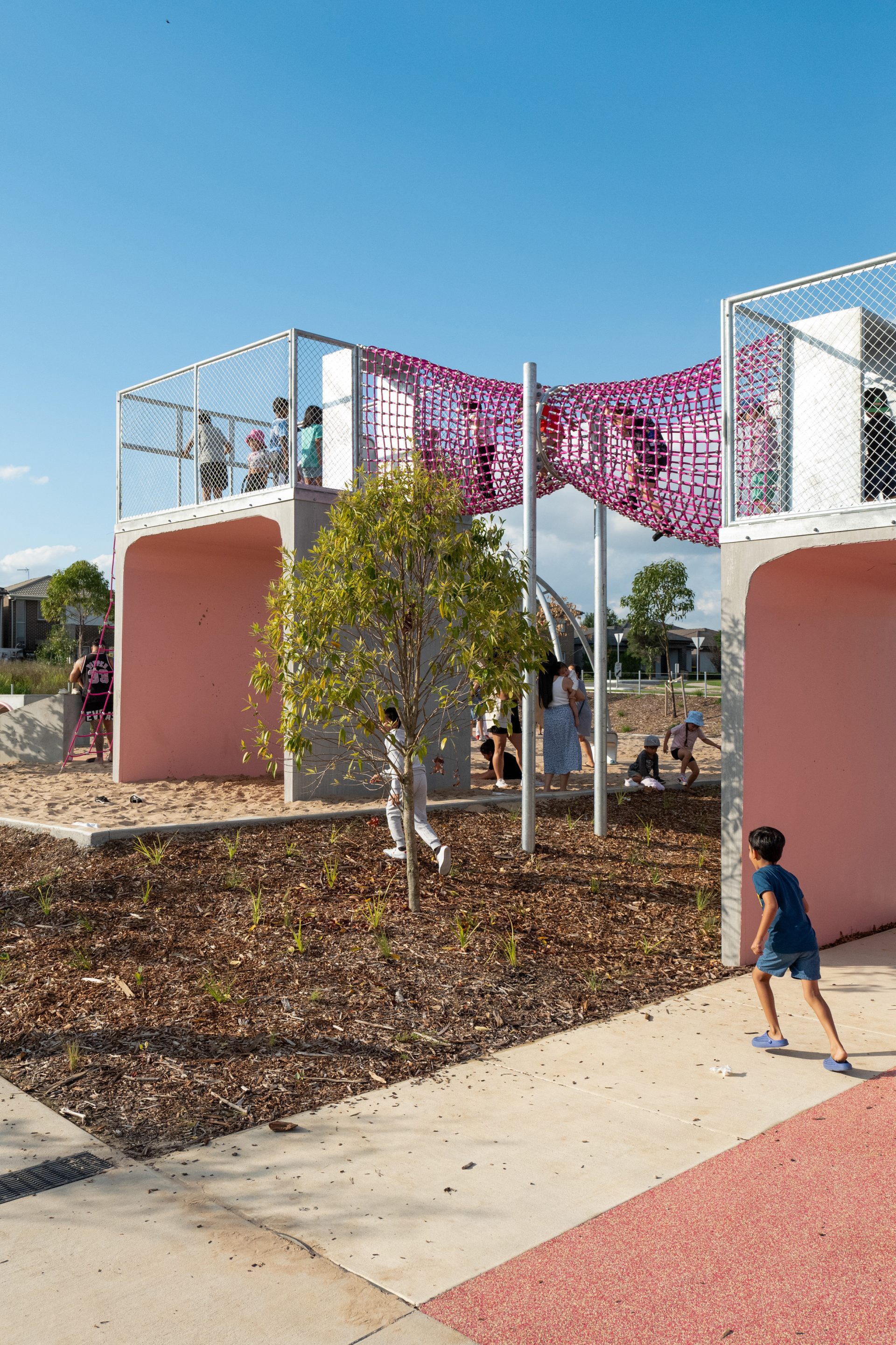 A delightful Galungara Park playground structure featuring pink climbing nets elevated above concrete platforms is set against a clear blue sky. Children are climbing and running around, and a small tree in the foreground is surrounded by mulch. A boy in a blue shirt dashes nearby.