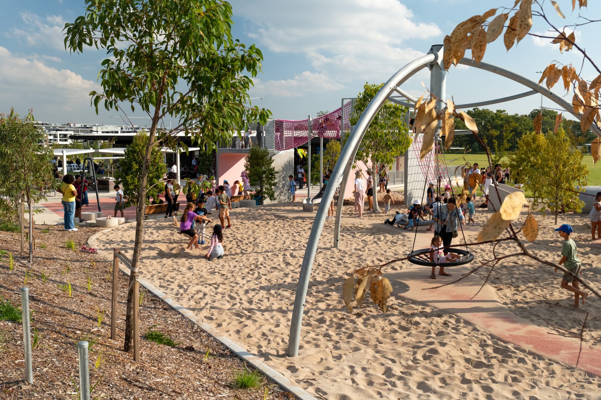 A lively Galungara playground with children playing on swings, climbing structures, and sand. Adults gather and watch from shaded areas. Trees and greenery surround the play area, set in a beautiful park under a blue sky with scattered clouds in the background.