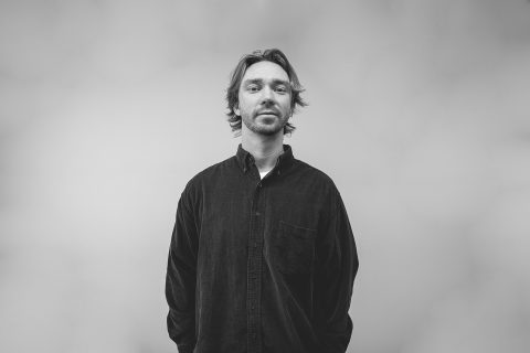 A person with medium-length hair stands against a plain background, wearing a dark button-up shirt. They are looking directly at the camera with a neutral expression. The image, capturing the essence of Sean Gyuris, is in black and white.