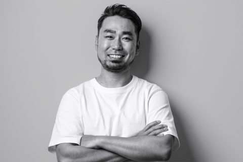 Black and white image of a man, Shota Saito, wearing a plain white t-shirt. He is smiling and has his arms crossed while standing against a plain background.