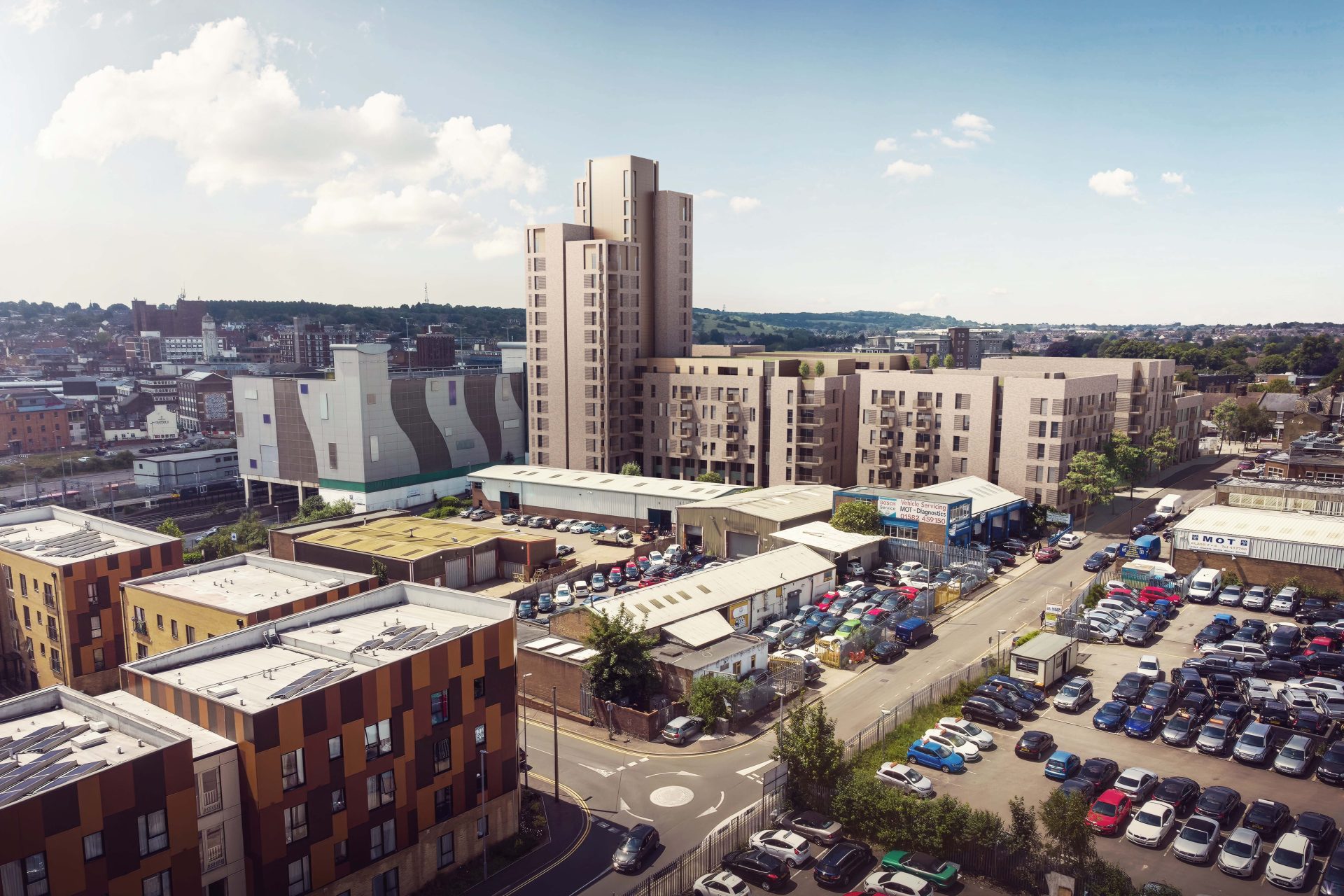 A cityscape featuring modern apartment buildings with a mix of architectural styles, centered around Station Place. A busy parking lot filled with cars is in the foreground, while the background shows additional urban buildings, green hills, and a partly cloudy sky.