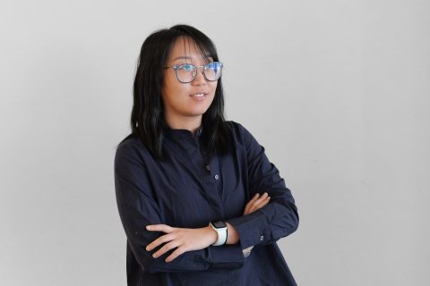 Sylvia Zhang, with long black hair and glasses, stands against a plain background in a dark long-sleeve shirt. Her arms are crossed as she looks slightly to the side with a neutral expression. A smartwatch adorns her left wrist.