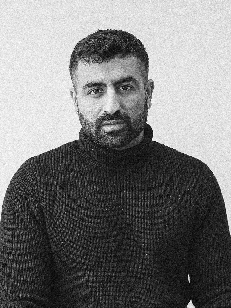 Black and white portrait of Tanuj Kalra with short, dark hair and a beard, wearing a black turtleneck sweater. He is facing the camera with a neutral expression against a plain, light-colored background.