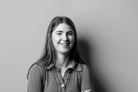 Black and white photo of a woman with long hair smiling at the camera. Tara Fischer is wearing a ribbed button-up top with a collar and has her arms crossed. The background is plain and minimalist.