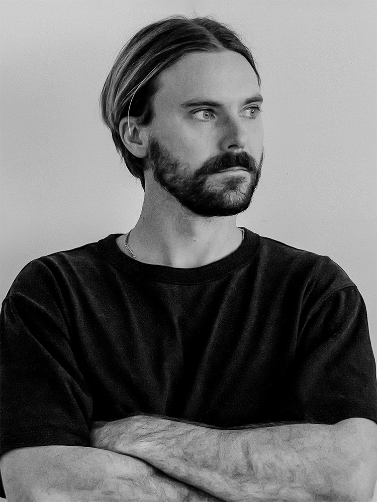 Black and white photo of Thomas Flugge, with shoulder-length hair and a beard. He is wearing a plain dark t-shirt and has his arms crossed, looking slightly to the right with a thoughtful expression. The background is plain and light-colored.
