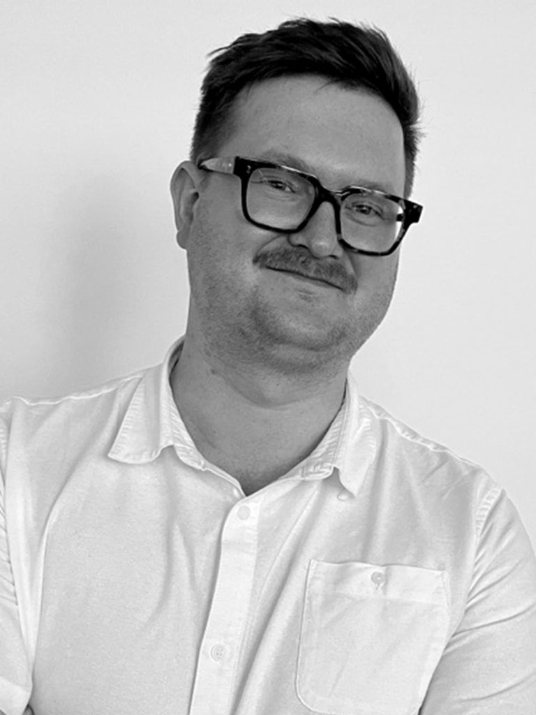 Black and white portrait of Thomas Chapman, a man with short hair, wearing glasses, a mustache, and a white button-up shirt with a pocket. He is smiling and looking at the camera with his arms casually crossed. The background is a plain light-colored wall.
