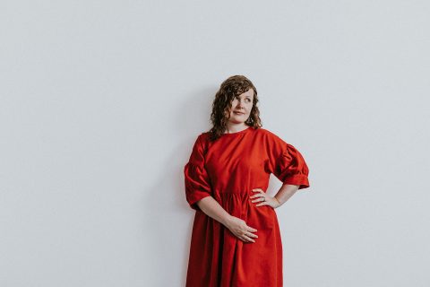 A woman with curly hair is standing against a plain white background. She is wearing a bright red, long-sleeved dress with puffed sleeves. One hand is on her hip, and the other hand rests gently on her abdomen as she looks slightly to her left, reminiscent of the elegance of Úna Breathnach-Hifearnáin.