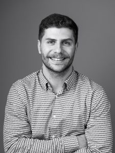 Black and white image of Xavi Font Sala smiling. He has short dark hair, light facial hair, and is wearing a patterned shirt. His arms are crossed in front of his chest, and he is standing against a plain background.