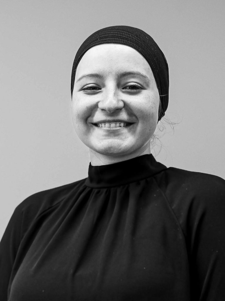 A black and white image shows a smiling woman, Yara Bassam, wearing a dark headscarf and a high-necked, long-sleeved top. She is looking directly at the camera, her face lit from the front, with a plain background.