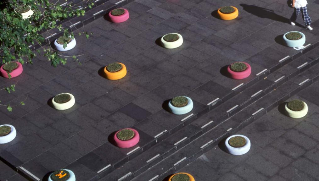 Aerial view of a paved area featuring colorful circular planters, reminiscent of amoebas, scattered across the pavement. The planters are in shades of yellow, orange, pink, and white. A person is walking near the planters, casting a long shadow on the ground. A tree with green leaves is partially visible.