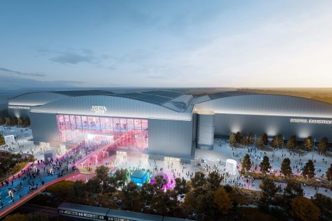 A large, modern exhibition center with the sign "Bristol Exhibition Centre" and the adjacent YTL Arena is bustling with people. The building features extensive glass windows and vibrant lighting at dusk. Visitors are gathered outside, and colorful lights illuminate the walkway.