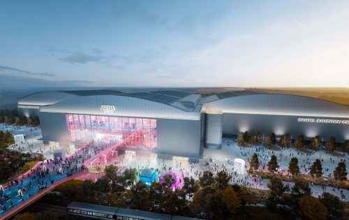 A large, modern exhibition center with the sign "Bristol Exhibition Centre" and the adjacent YTL Arena is bustling with people. The building features extensive glass windows and vibrant lighting at dusk. Visitors are gathered outside, and colorful lights illuminate the walkway.