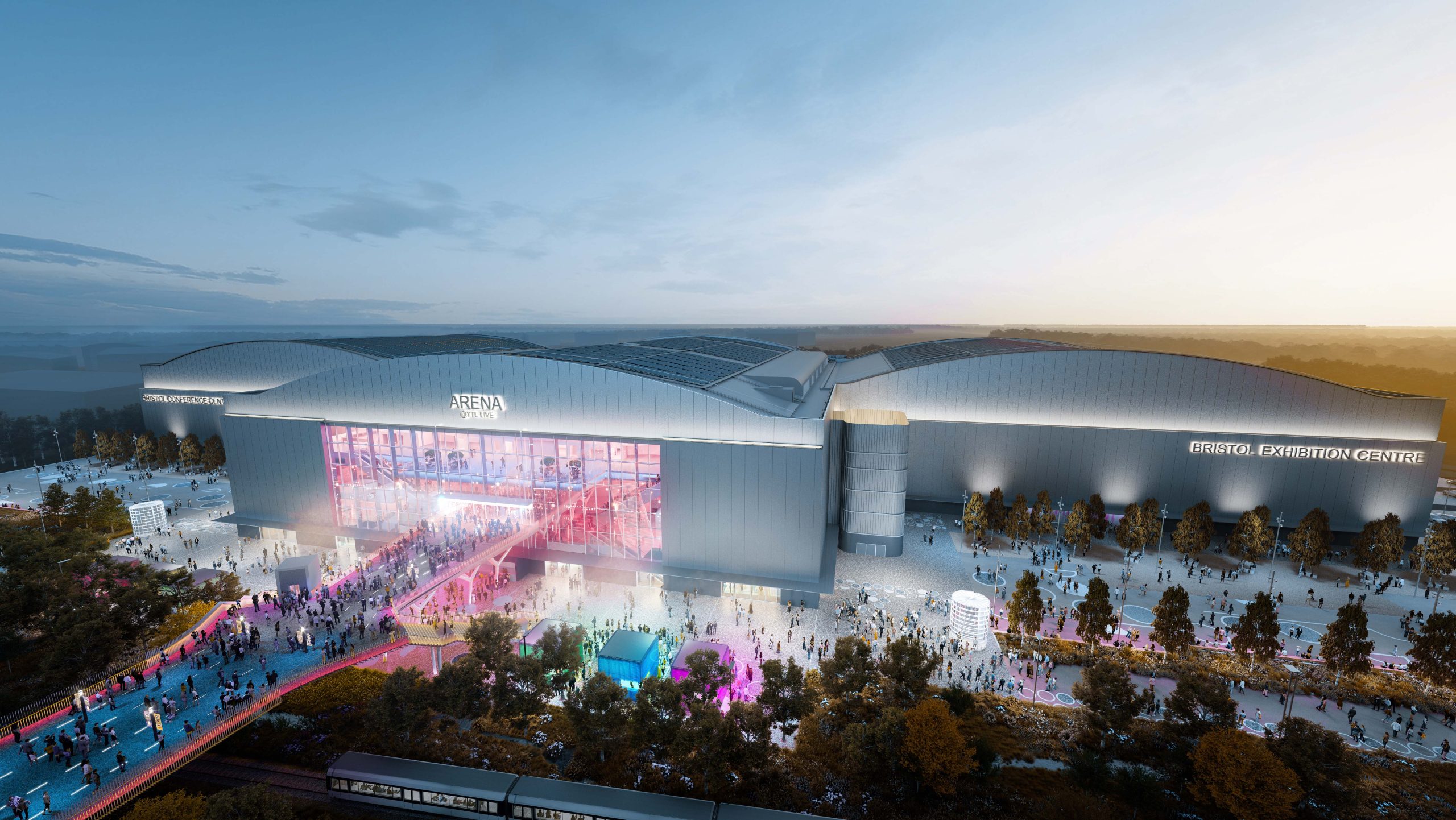 A large, modern exhibition center with the sign "Bristol Exhibition Centre" and the adjacent YTL Arena is bustling with people. The building features extensive glass windows and vibrant lighting at dusk. Visitors are gathered outside, and colorful lights illuminate the walkway.