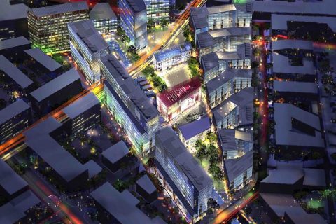 Aerial night view of a cityscape illuminated with colorful lights. Various modern buildings with glass facades cluster around Green Square, surrounded by interconnected streets and trees. The vibrant lighting highlights the urban architecture and street patterns in the bustling town center.