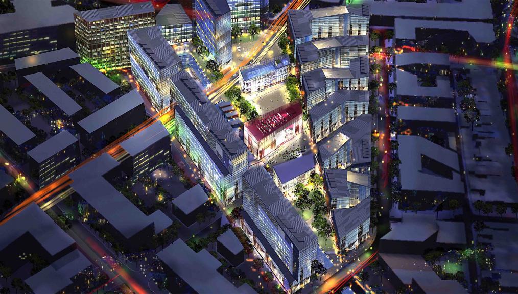 Aerial night view of a cityscape illuminated with colorful lights. Various modern buildings with glass facades cluster around Green Square, surrounded by interconnected streets and trees. The vibrant lighting highlights the urban architecture and street patterns in the bustling town center.