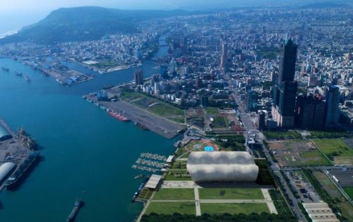 Aerial view of Kaohsiung, a coastal city with a prominent river running through it. The vibrant cityscape features numerous high-rise buildings, a bustling port with ships docked, and a large, bubble-like stadium near a green park area, close to the Convention Centre hosting various exhibitions.