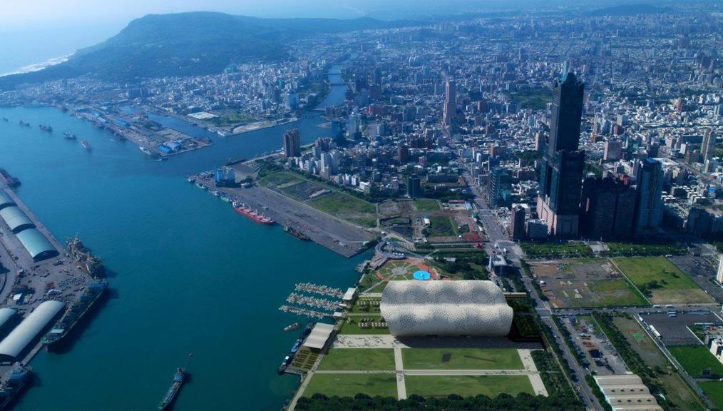 Aerial view of Kaohsiung, a coastal city with a prominent river running through it. The vibrant cityscape features numerous high-rise buildings, a bustling port with ships docked, and a large, bubble-like stadium near a green park area, close to the Convention Centre hosting various exhibitions.