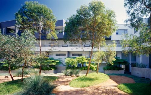 Modern NOVA apartment complex with large windows and balconies, surrounded by lush green trees and plants in a well-maintained garden with curving walkways. Bright, sunny day enhancing the vibrant greenery and contemporary architecture.