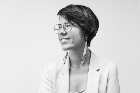 A person with short hair, wearing glasses and long earrings, is smiling while looking to the side. Dressed in a light-colored blazer over a top, the individual exudes effortless style. The plain white background enhances the minimalistic feel of this captivating portrait by Daria Vazhenina.