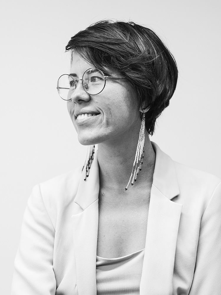 Black and white portrait of Daria Vazhenina, a person with short hair and glasses, smiling and looking to the side. They are wearing a blazer and long, beaded earrings. The background is plain, drawing focus to their expression and attire.