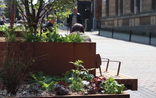 A person sits on a wooden bench next to a planting bed filled with various green and red-leafed plants in an urban setting. A tree and additional plants are also visible in the planter. The background shows Gloucester’s historic buildings with a few people walking through this vibrant heritage action zone.