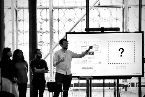 A group of four people stand in front of a large screen during a presentation at Melbourne Design Week. One person, a man in a white shirt, gestures toward the screen displaying a large question mark. The background features a wall with circular patterns and large windows. The mood appears collaborative.